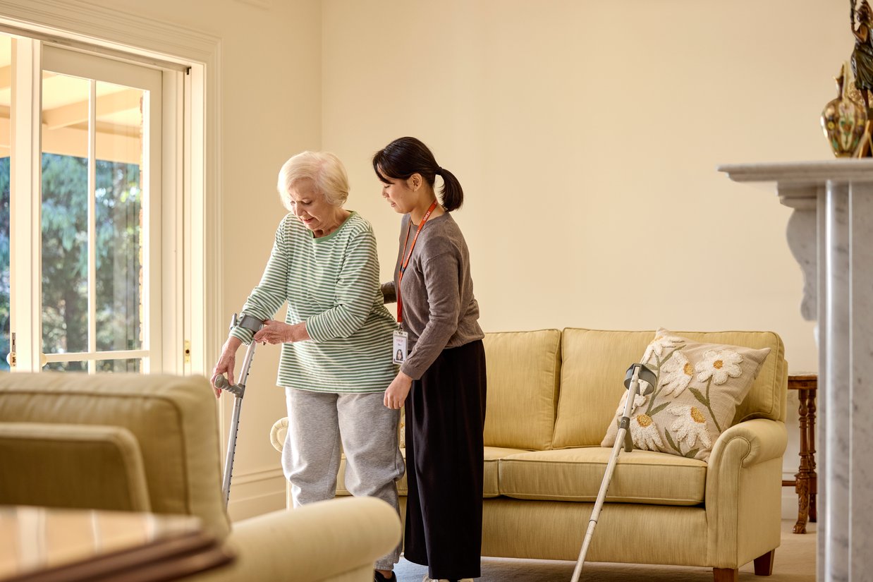 A young woman helps an older women on crutches off of the couch.