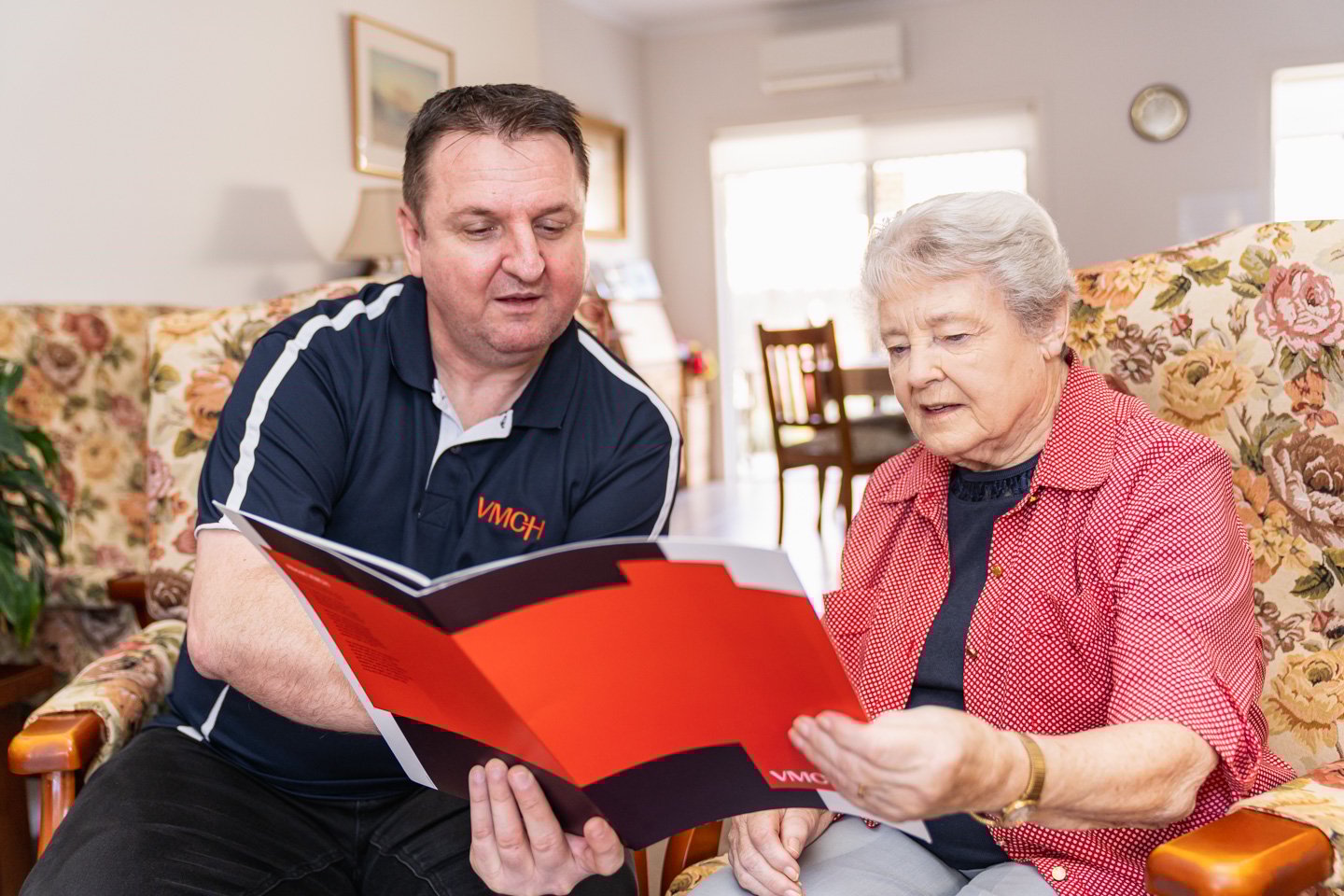 A VMCH care partner sitting with an older lady on a couch looking at a folder of information