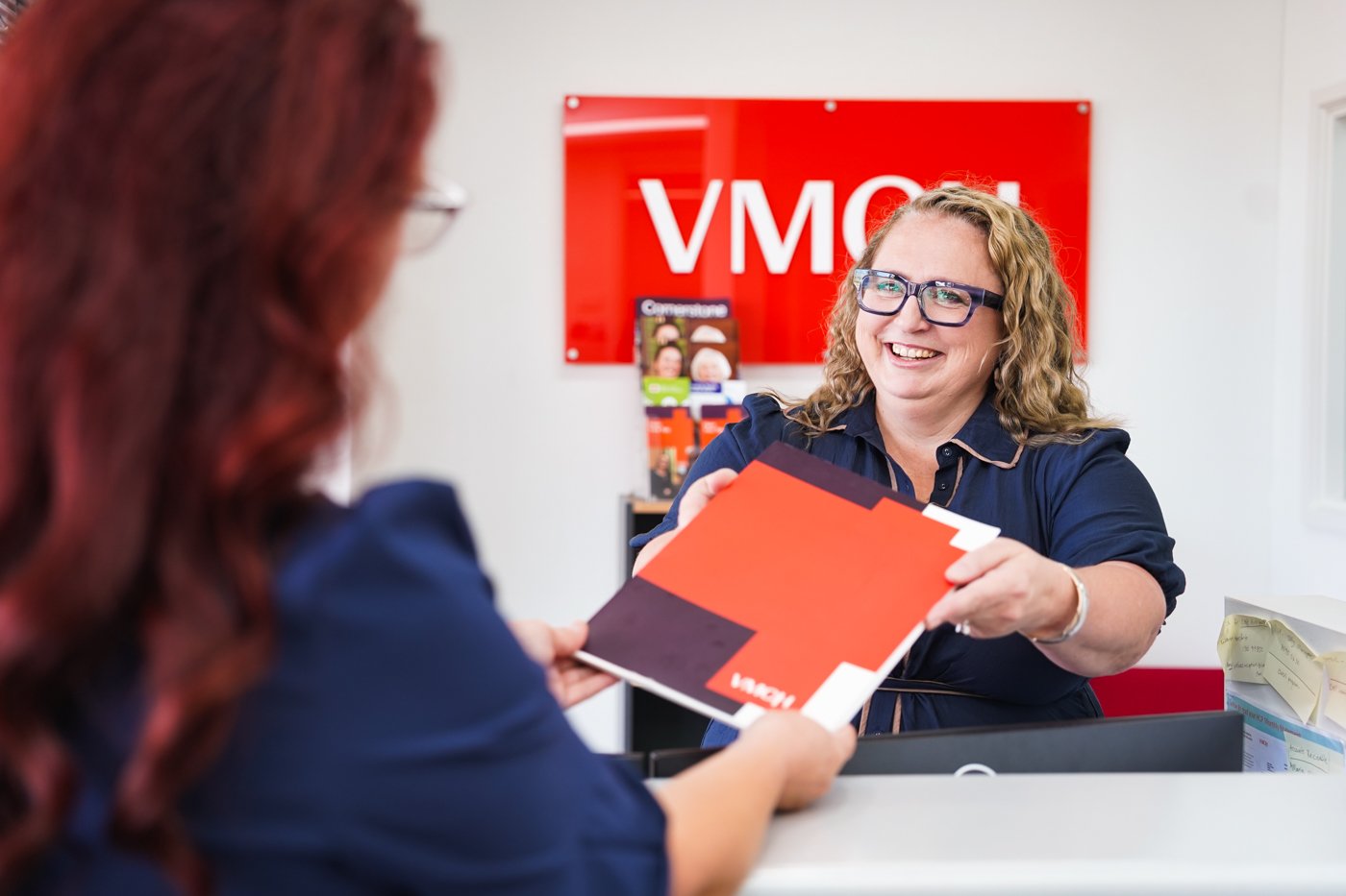 A woman sitting behind a shop counter handing a brochure to another woman.