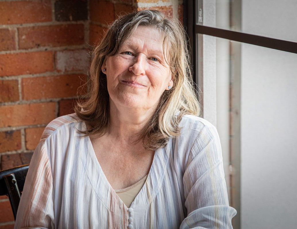 A woman smiling warmly, sitting by a window.