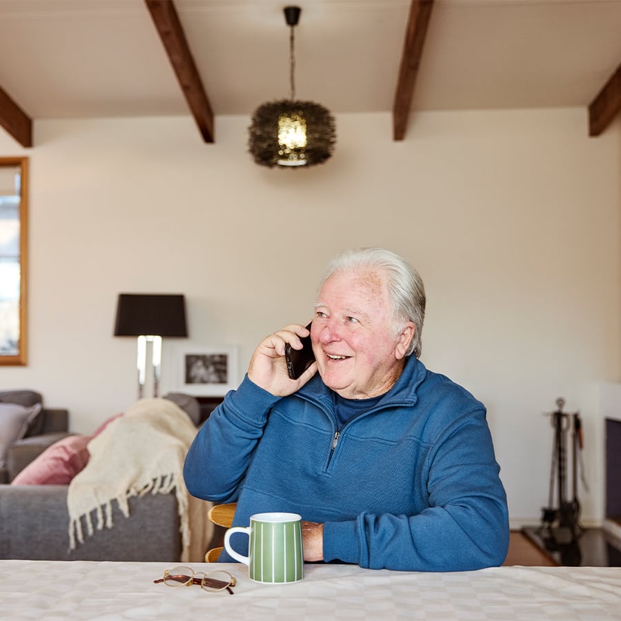 VMCH aged care resident sitting at a table with a coffee, smiling while talking on the phone at home.