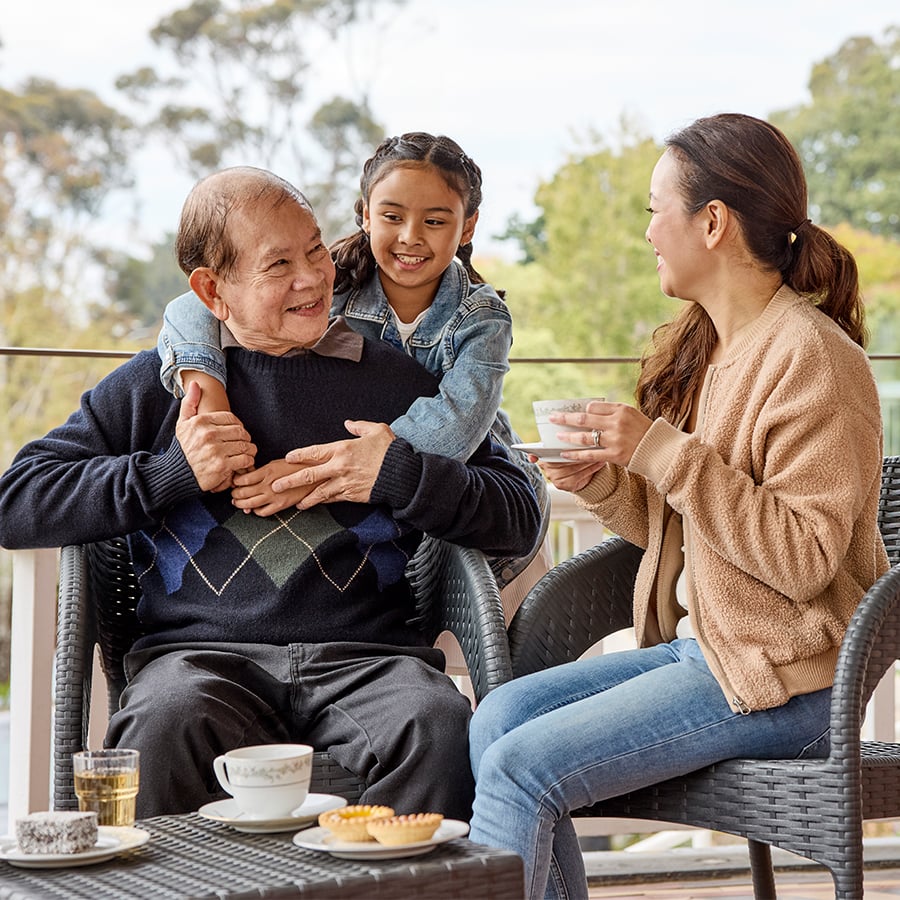 VMCH aged care resident enjoying family time on the balcony, smiling with his daughter and granddaughter over tea and conversation.