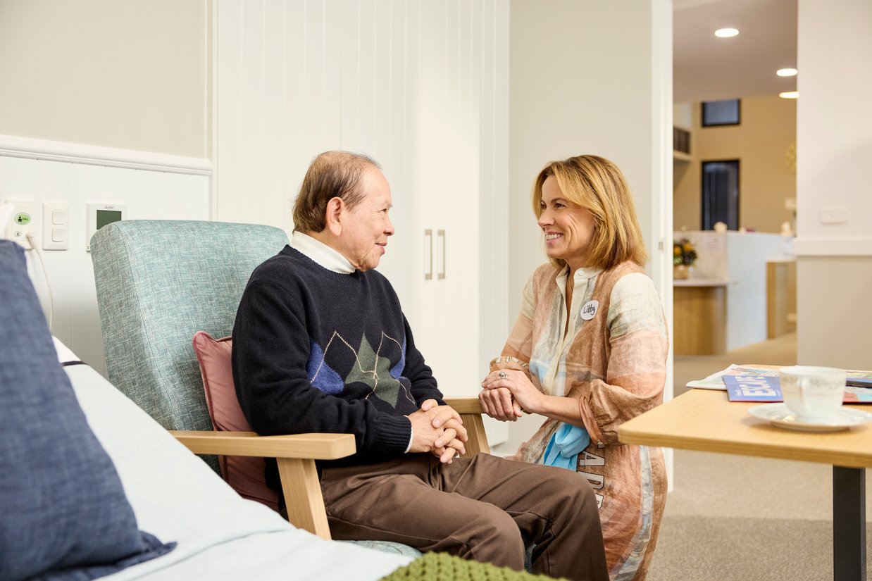 An elderly man sitting in an armchair with a VMCH pastoral care worker smiling and talking with him