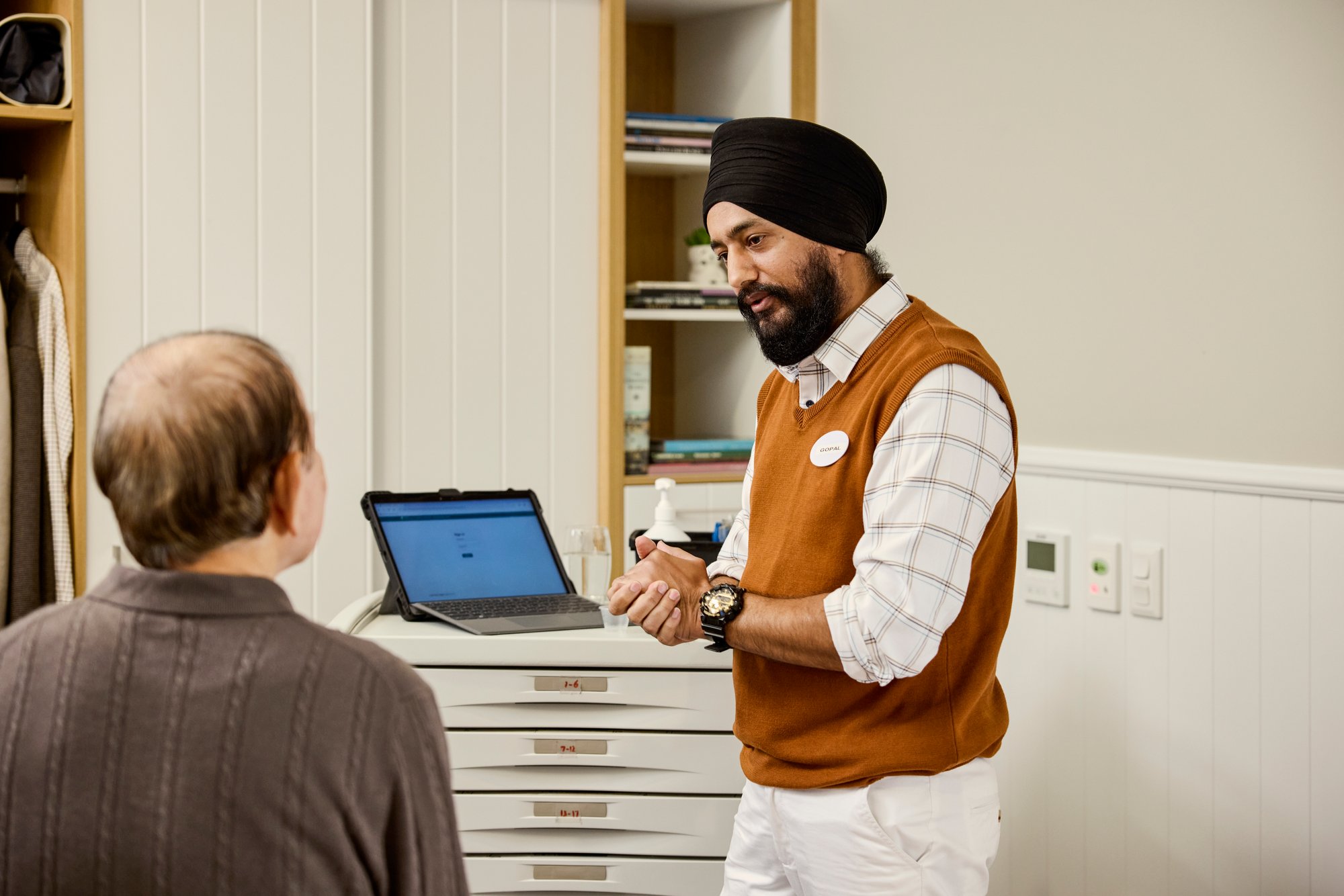 An Indian male nurse is talking with an elderly aged care resident with a laptop in the background