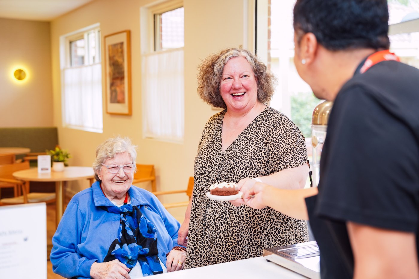 A woman being handed a piece of cake in a cafe, with an older lady smiling behind her.