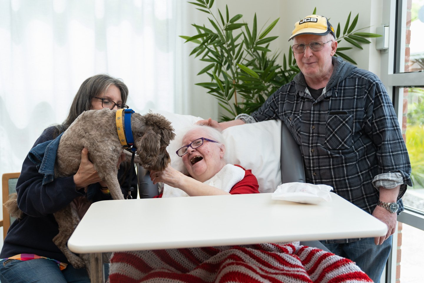 An elderly woman in a bed with an elderly man by her right hand side. A woman with a grey therapy dog are to the woman's left