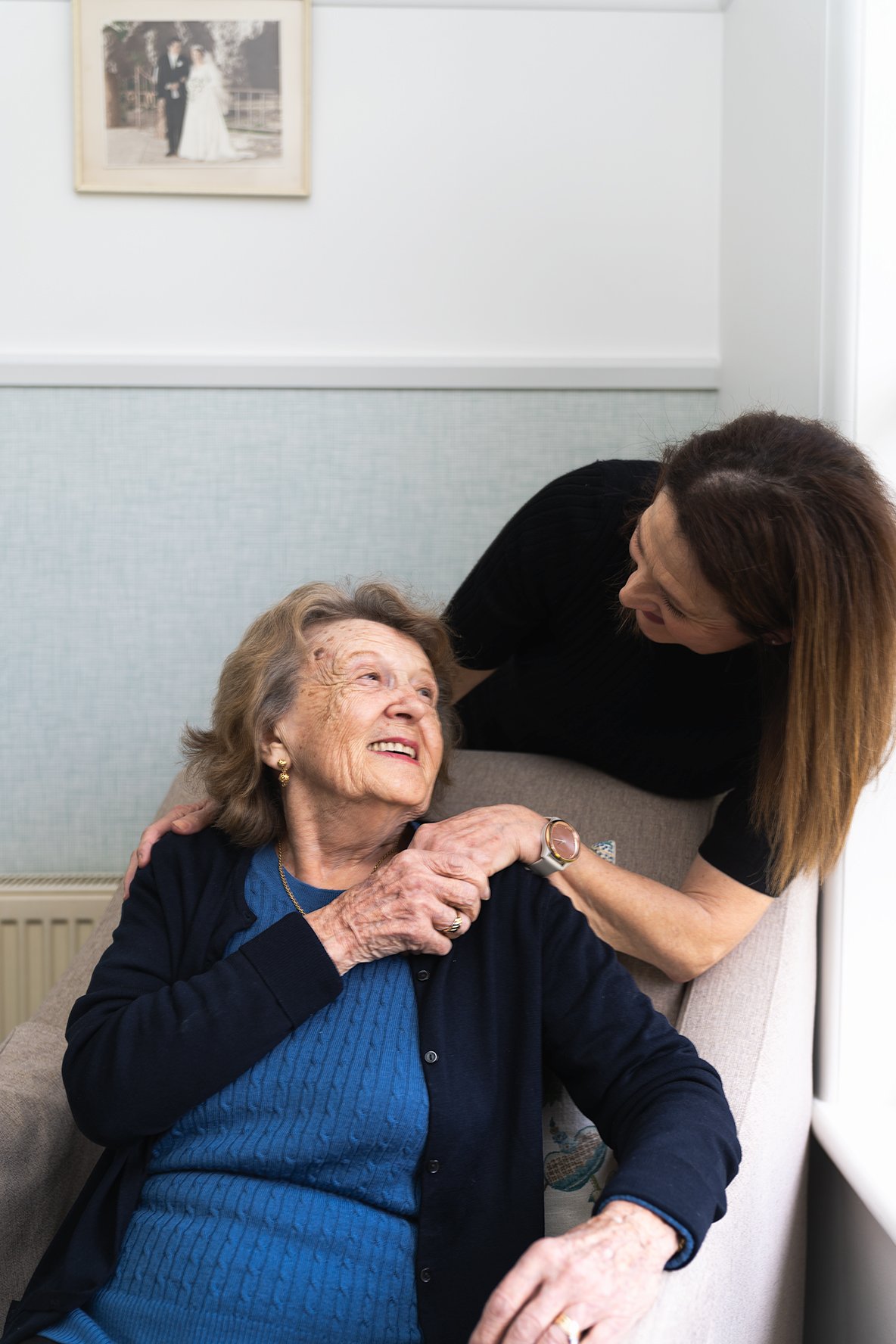 An elderly woman sitting in a chair with her daughter looking over her shoulder and holding her hand