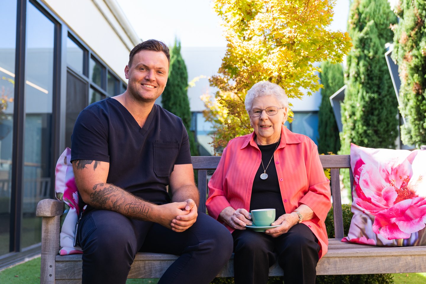 A male aged care worker sitting with an elderly woman outside on a bench. She is drinking a cup of tea